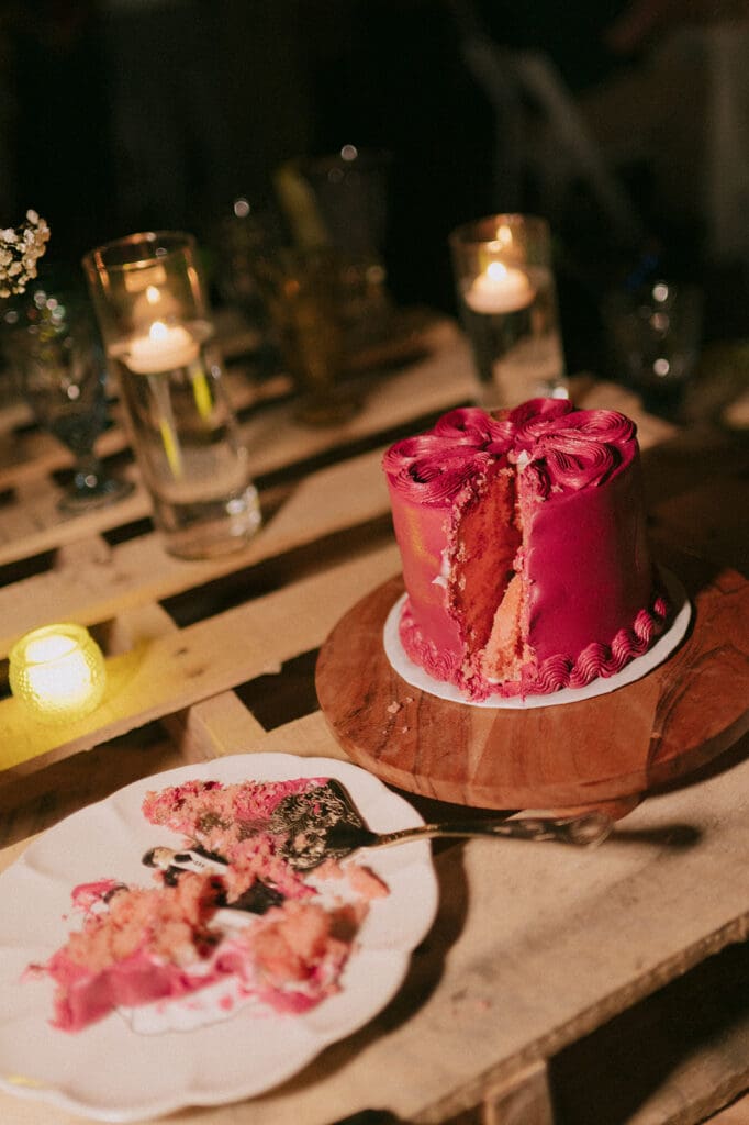 A partially cut pink frosted cake sits on a wooden board, with several slices on a white plate nearby; candles and glasses are in the background.