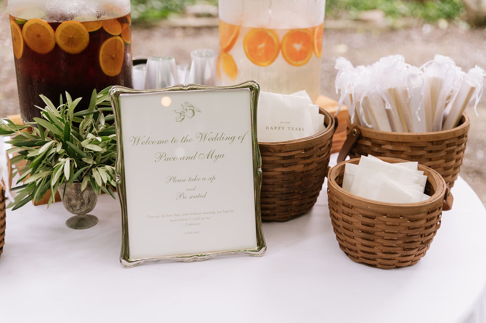 A round table with drinks and snacks sits under a white umbrella in a wooded outdoor setting with tall trees and green foliage.