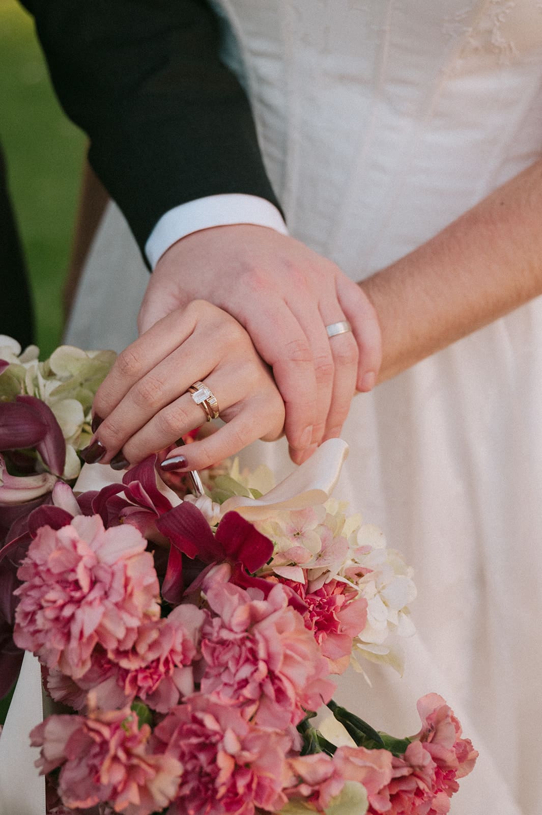 Close-up of a couple’s hands with wedding rings resting on a bouquet of pink and burgundy flowers, with the bride in a white dress and the groom in a black suit.