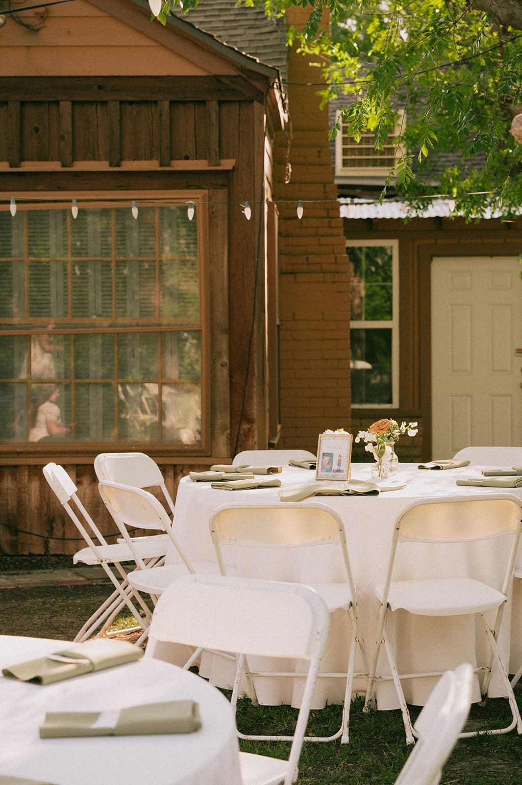 Round tables with white tablecloths and folding chairs set up outdoors near a wooden building, with plates, napkins, and simple floral centerpieces on the tables at a Small Wedding Venues in Dallas, TX 
