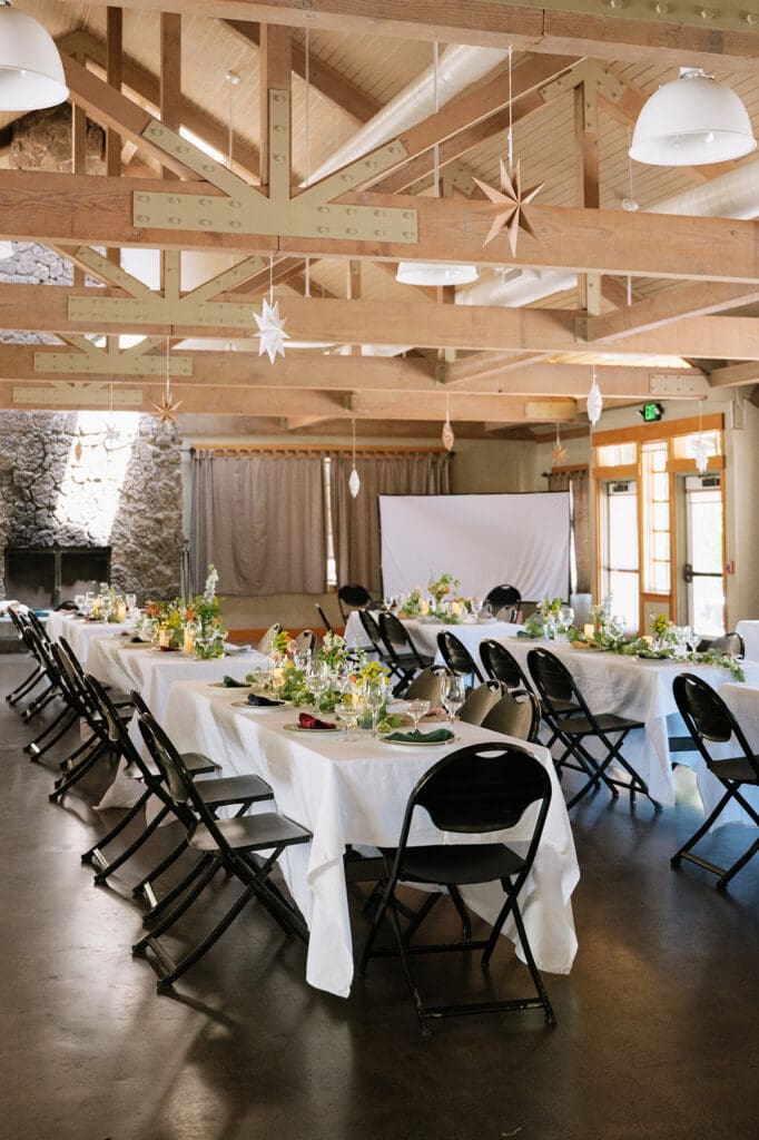 Tables with white tablecloths and black chairs are set for an event in a rustic room with wooden beams and natural light | 9 Wedding Venues in Oregon for an Intimate Celebration
