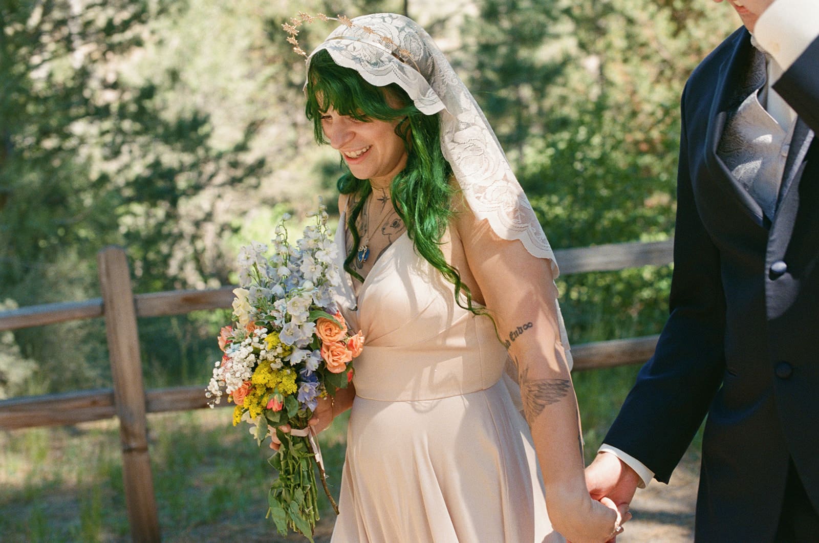 A bride in a cream dress and veil, holding a bouquet, walks outdoors with a person in a suit. Trees and a wooden fence are visible in the background.