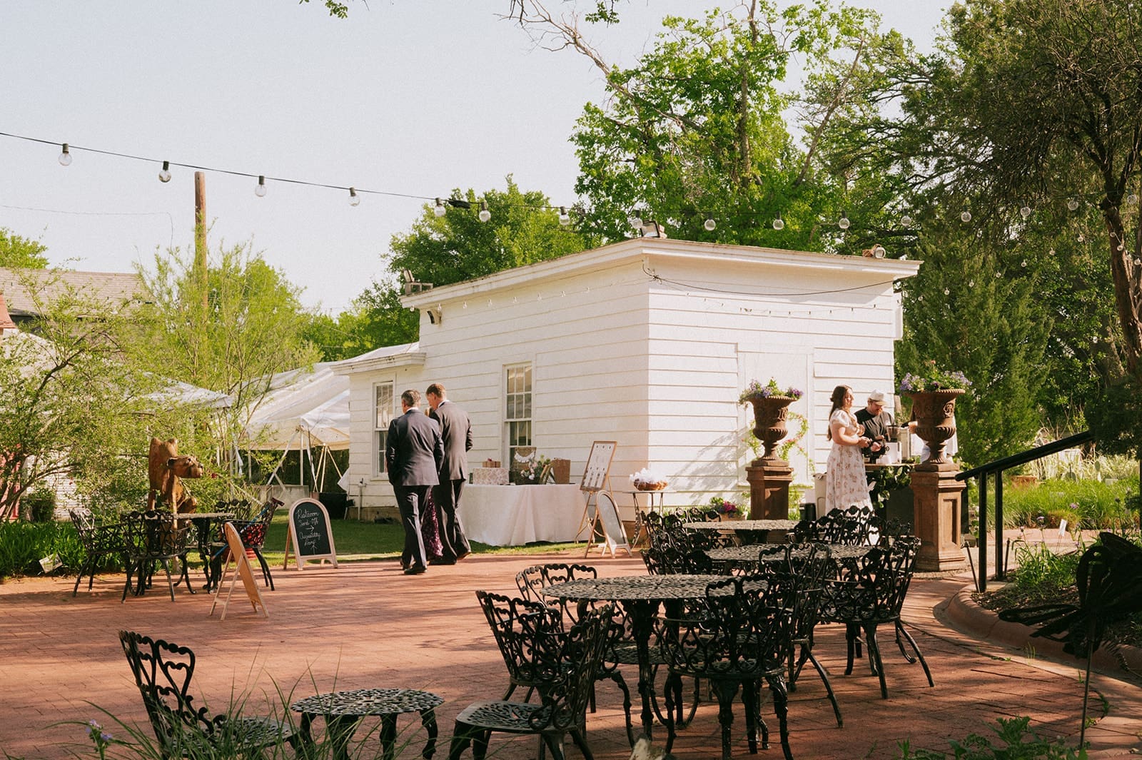 Outdoor event with empty wrought iron tables and chairs, a few people in formal attire standing near a white building, surrounded by greenery and string lights above.