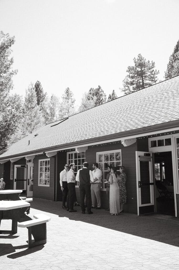 A group of people stands and talks outside a building with open doors, surrounded by trees; picnic tables are in the foreground | 9 Wedding Venues in Oregon for an Intimate Celebration