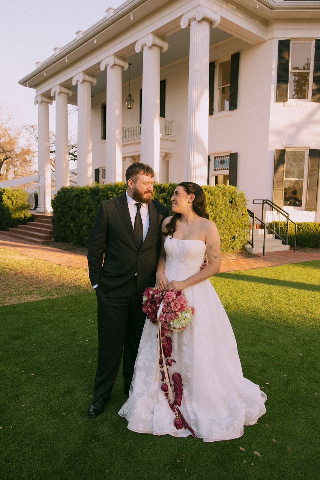 A bride and groom stand together on a lawn in front of a large white building with columns, smiling at each other.