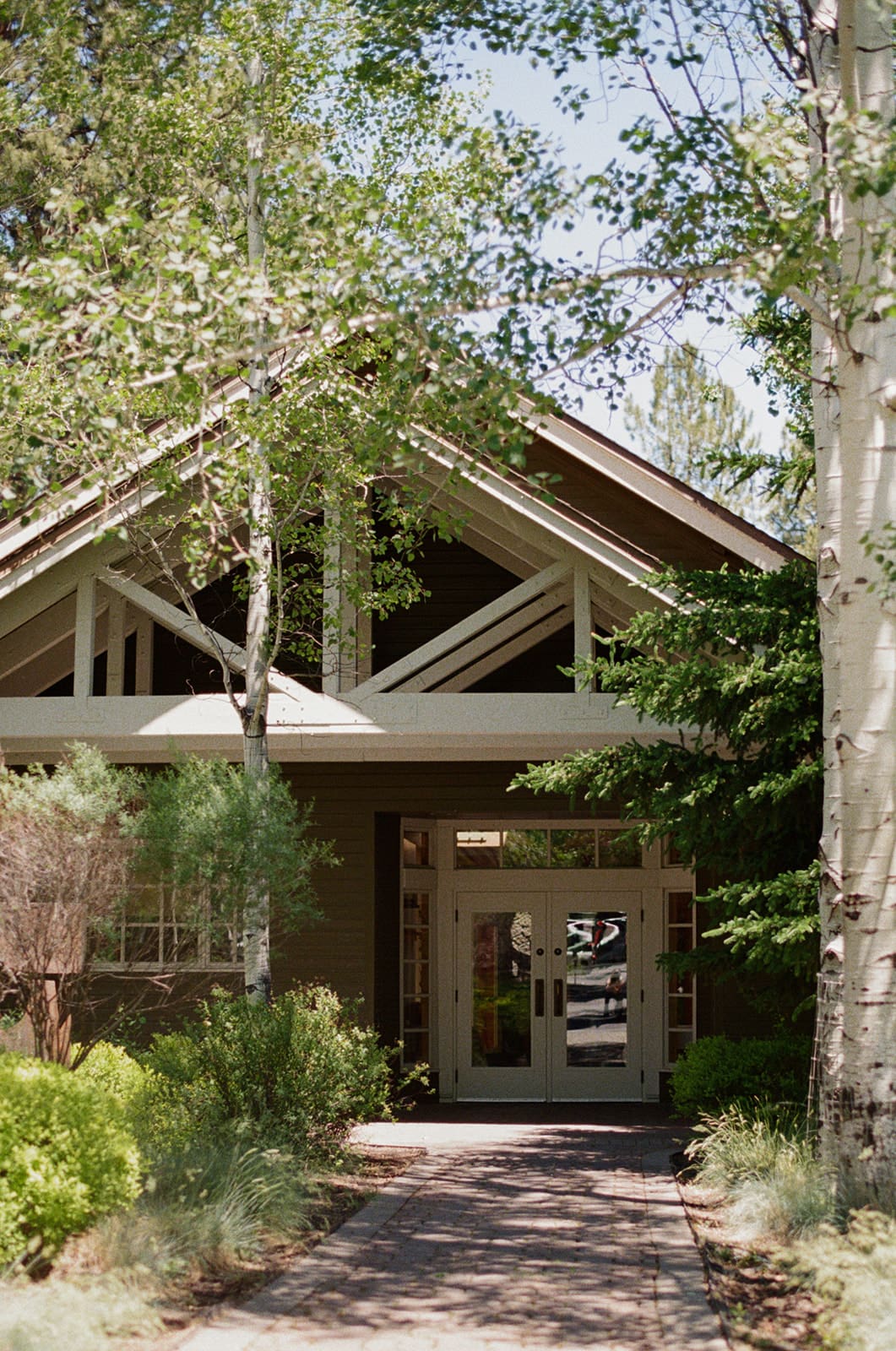 Modern house exterior with large windows, a peaked roof, and a pathway leading to the front door, surrounded by trees and greenery.
