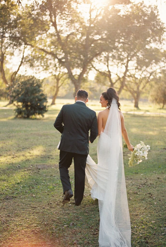 Bride in a white dress and veil walks hand in hand with a man in a suit through a sunlit outdoor setting with trees in the background.