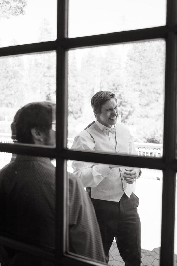 Two men are standing outside, viewed through window panes. One is dressed in formal attire and smiling, while the other is partially visible. Trees and outdoor scenery are in the background.