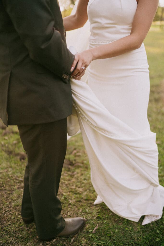Bride in a white dress and veil walks hand in hand with a man in a suit through a sunlit outdoor setting with trees in the background.