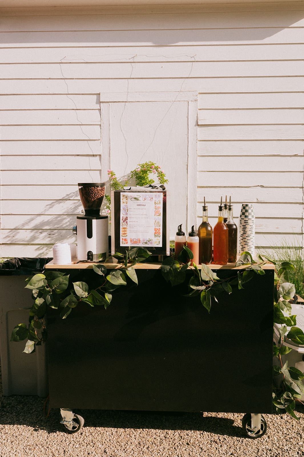 A small drink station on wheels is decorated with greenery and bottles, set against a white wooden wall in outdoor daylight.
