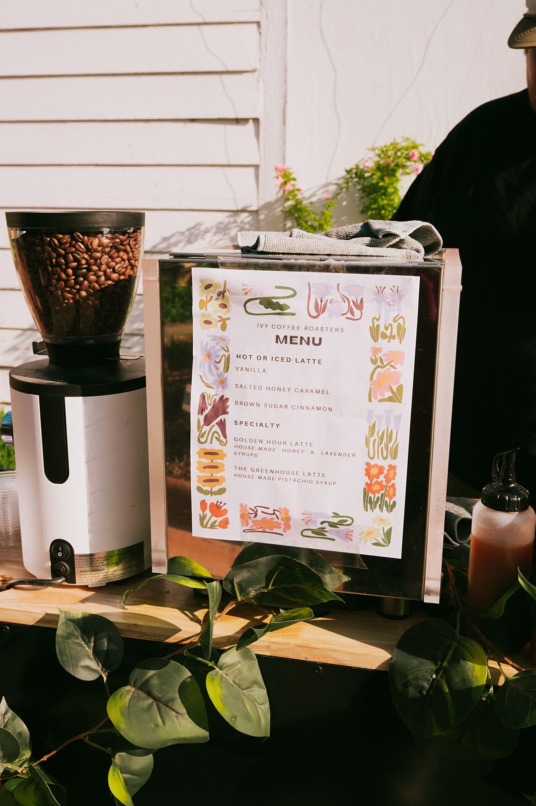 A small drink station on wheels is decorated with greenery and bottles, set against a white wooden wall in outdoor daylight.