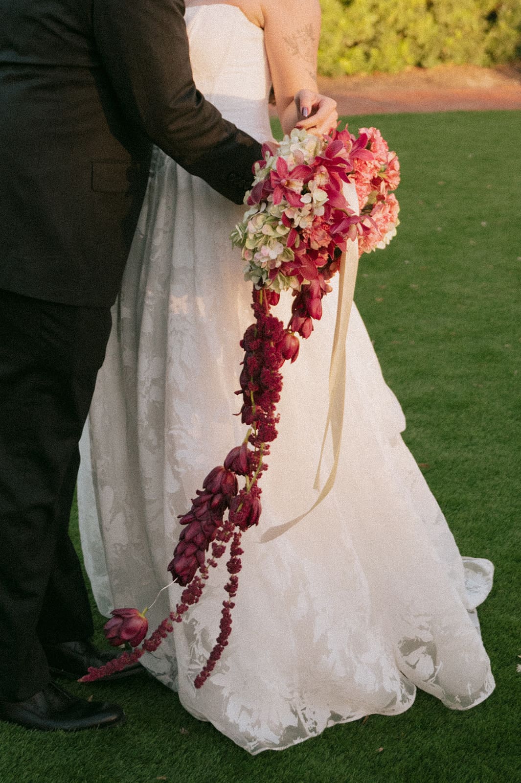 Close-up of a couple’s hands with wedding rings resting on a bouquet of pink and burgundy flowers, with the bride in a white dress and the groom in a black suit.
