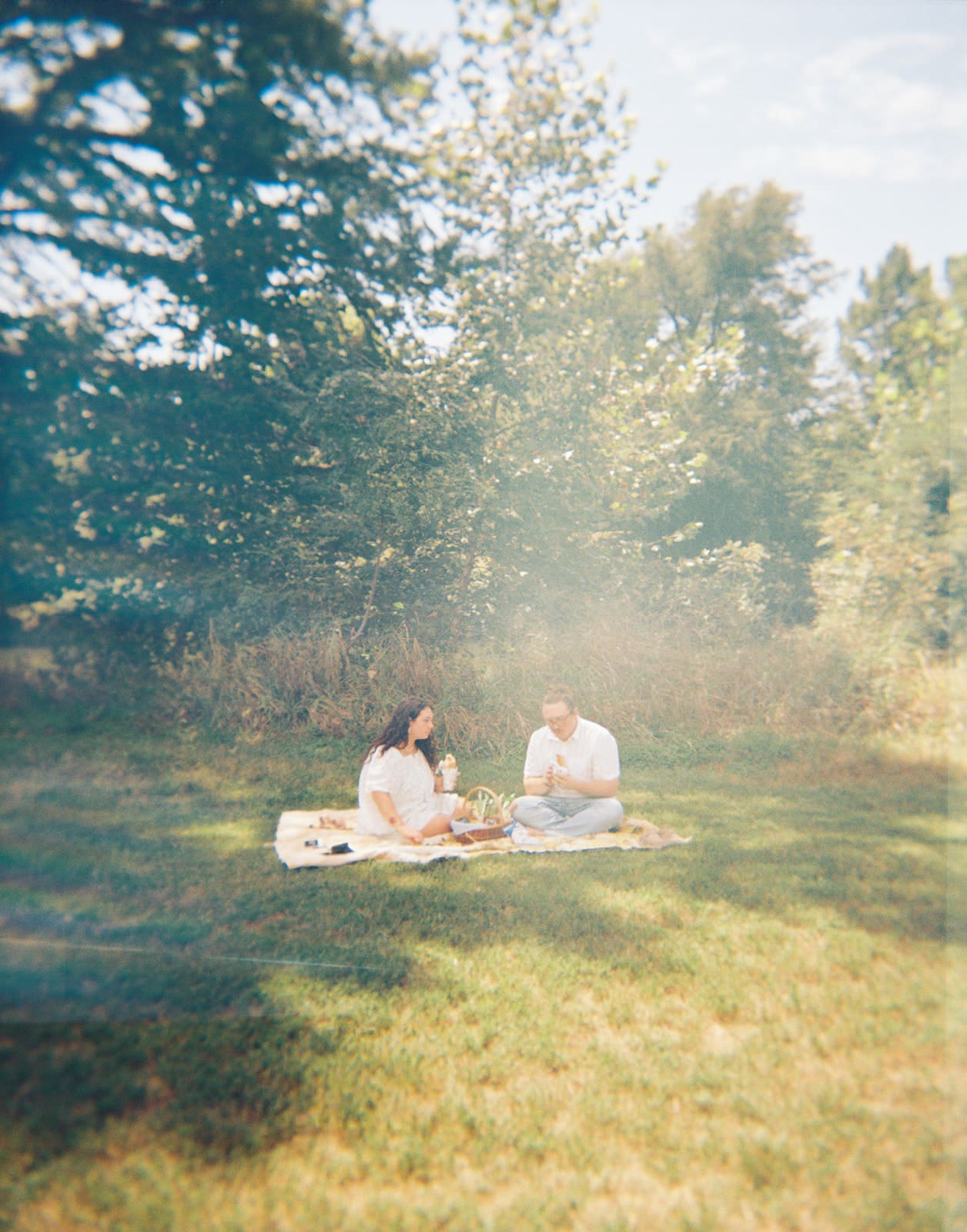 Two people stand in shallow water near a stone edge, holding hands and smiling at each other, with trees and a pond in the background.
