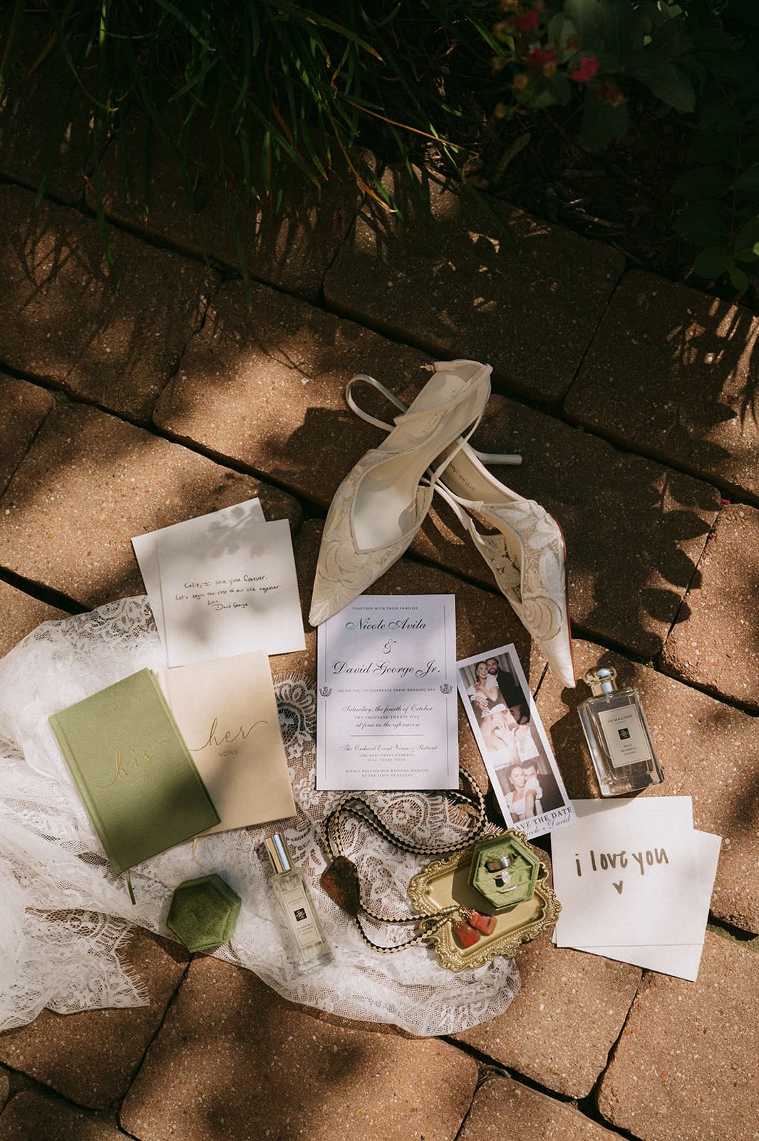 A flat lay of invitation cards, a green notebook, a small box, perfume bottles, a lace cloth, a ribboned pouch, and a photo on a stone surface with some foliage shadows.