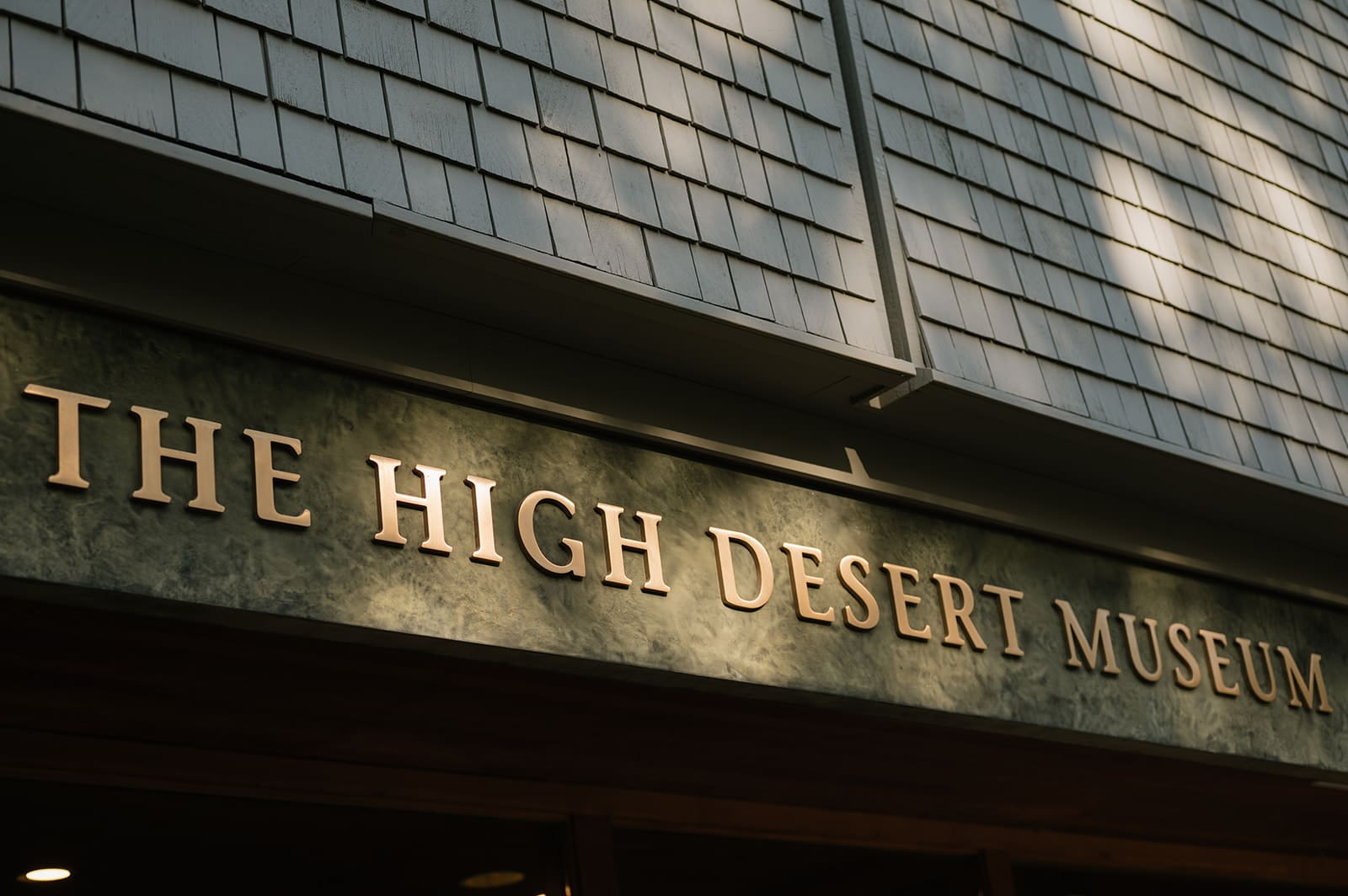 The exterior of a building with a sign that reads “The High Desert Museum” in large gold letters beneath grey shingle siding.