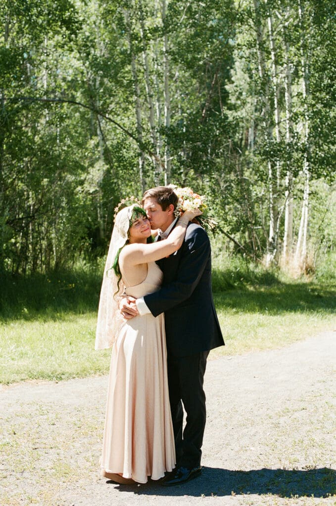 A bride and groom stand close together outdoors, smiling at each other. The bride holds a bouquet of flowers and wears a lace veil. Trees and a pond are visible in the background| 9 Wedding Venues in Oregon for an Intimate Celebration