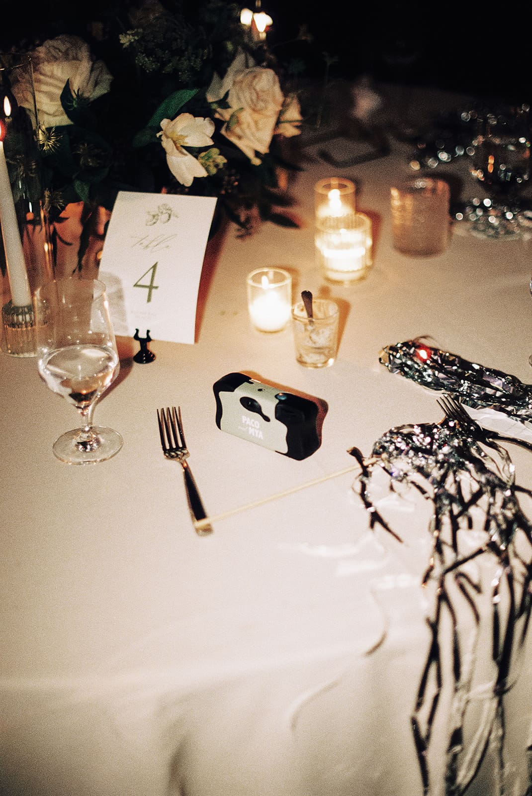 A round table set with a centerpiece, candles, a table number 4 card, a glass, cutlery, a disposable camera, and metallic party decorations. | Unique Wedding Ideas