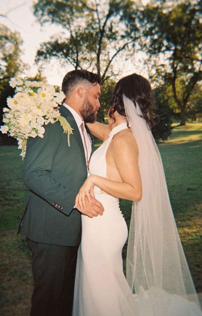 Bride in a white dress and veil walks hand in hand with a man in a suit through a sunlit outdoor setting with trees in the background.