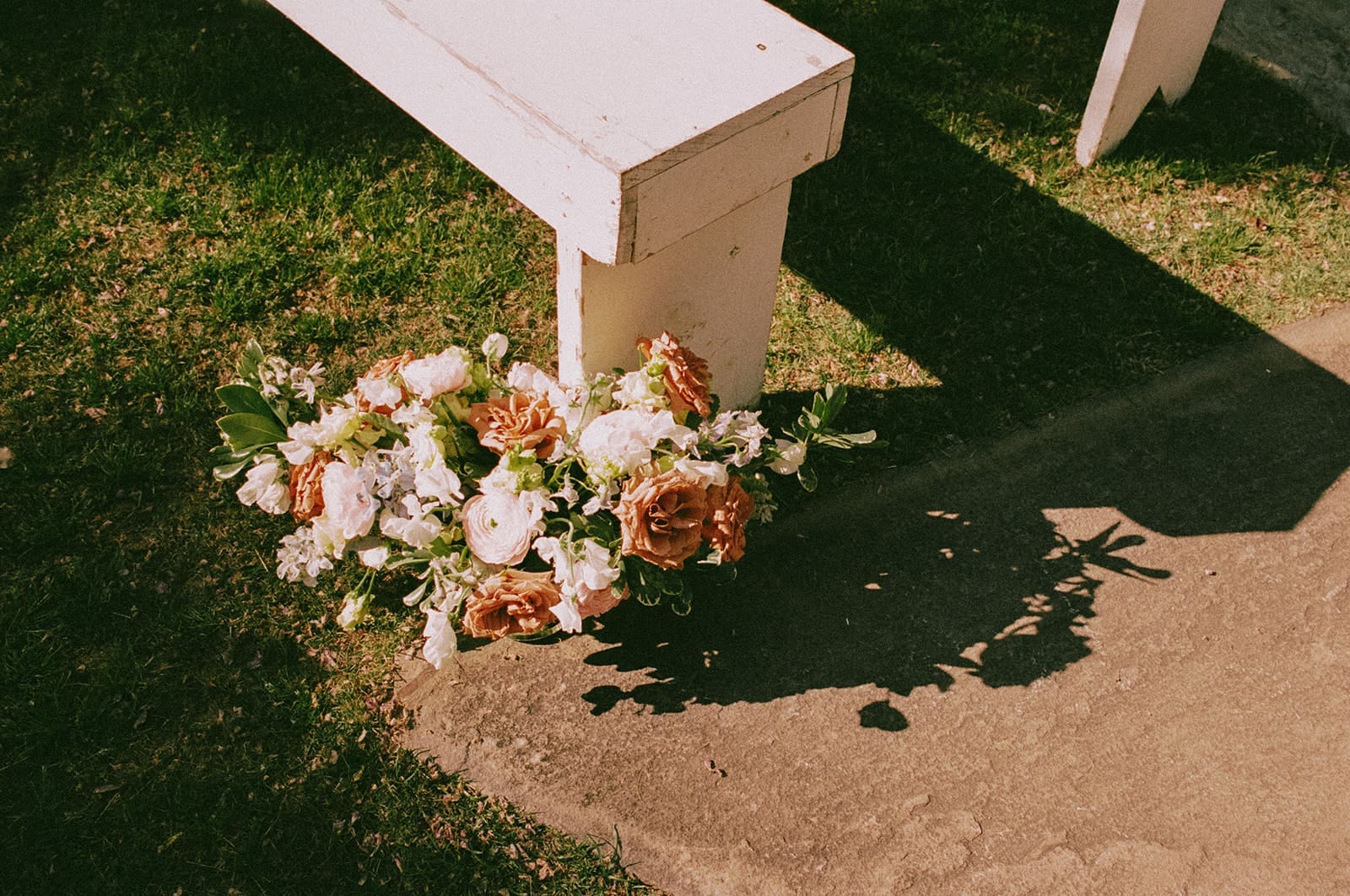 A bouquet of pink and white flowers lies on the ground next to a white wooden bench on a grassy area.