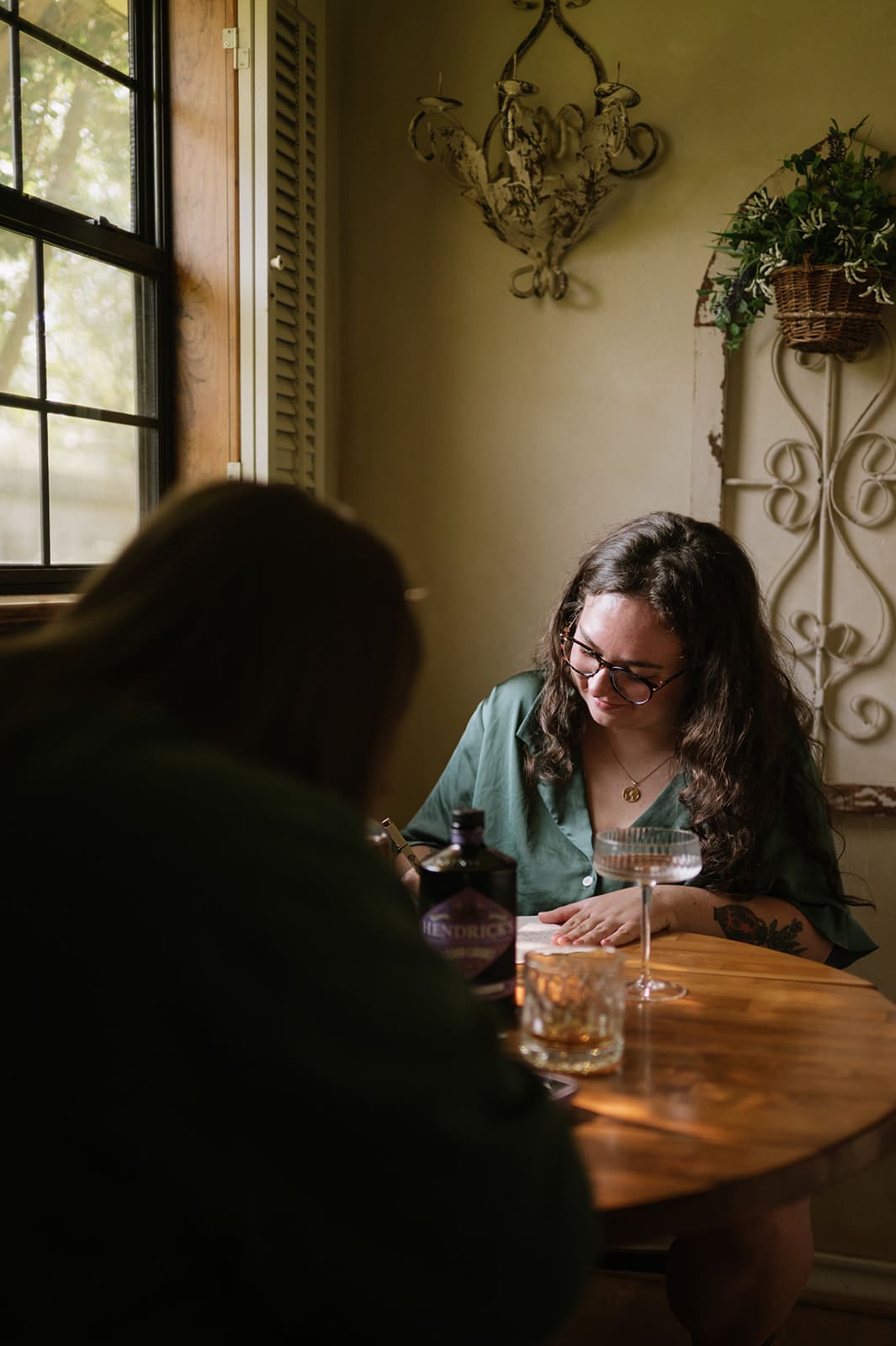 Two people sit at a wooden table by a window, with drinks and a bottle on the table. One person is smiling and looking down, while the other is facing away from the camera.