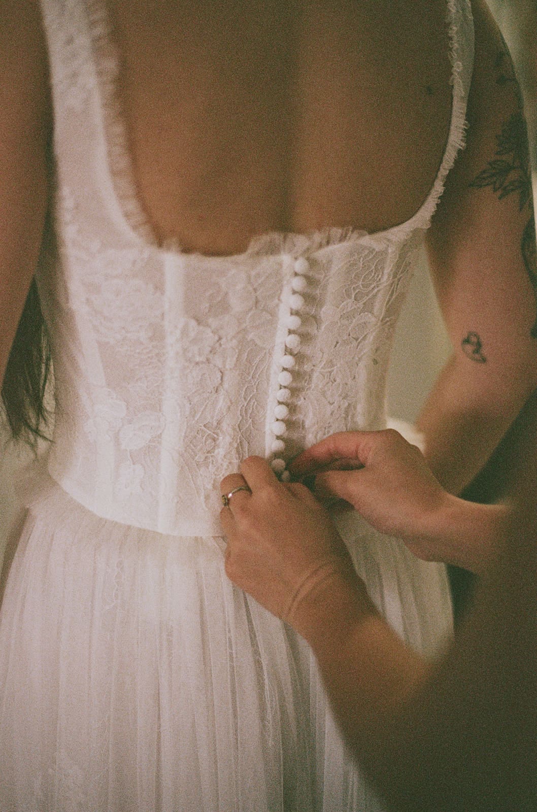 Hands are fastening buttons on the back of a white lace dress worn by a person with bare shoulders.