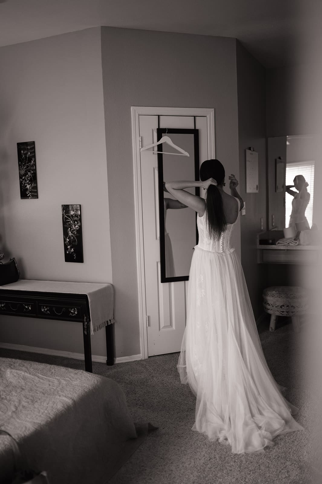 A woman in a white dress stands in a bedroom, facing a mirror and holding a hanger above her head, with her reflection visible in another mirror. | Unique Wedding Ideas