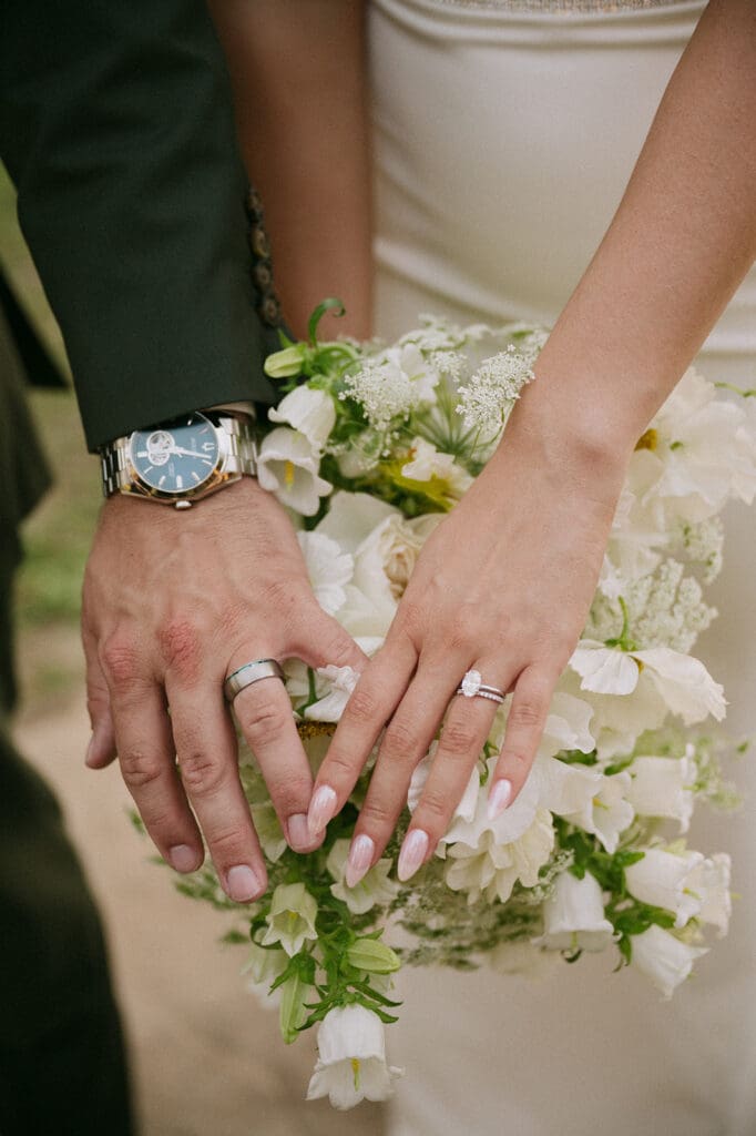 Bride in a white dress and veil walks hand in hand with a man in a suit through a sunlit outdoor setting with trees in the background.