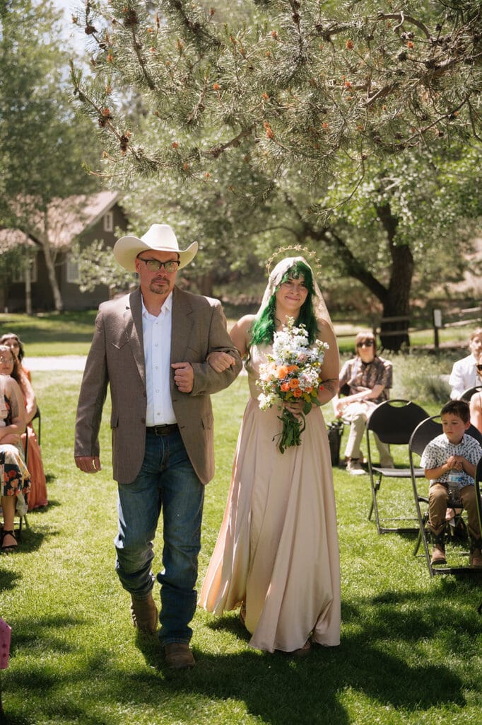 A person in a cowboy hat walks beside a person in a long dress holding a bouquet at an outdoor ceremony with seated guests | 9 Wedding Venues in Oregon for an Intimate Celebration
