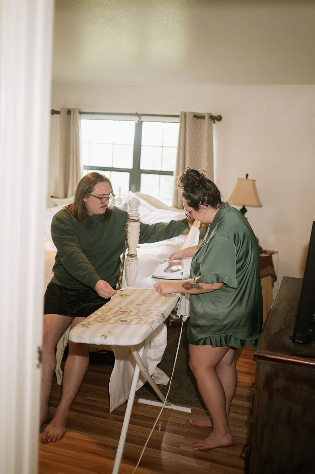 Two people are in a bedroom; one is sitting on a bed while the other stands at an ironing board, appearing to iron clothes.
