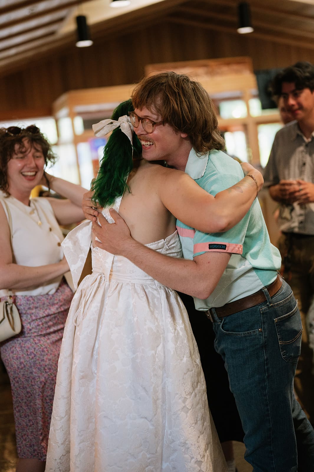 A person in a white dress hugs another person in a light blue shirt at an indoor gathering, while others stand nearby smiling.