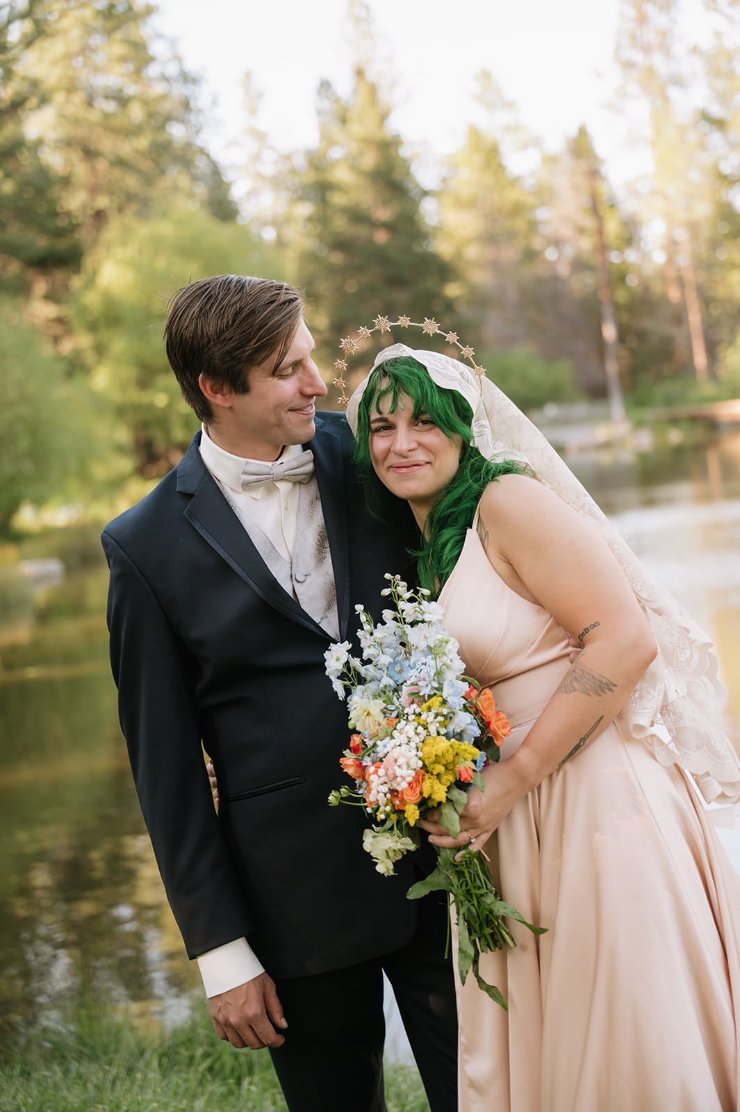 A couple poses outdoors by a lake; the woman in a light dress holds a bouquet and has green hair, while the man in a suit stands behind her, both looking happy.