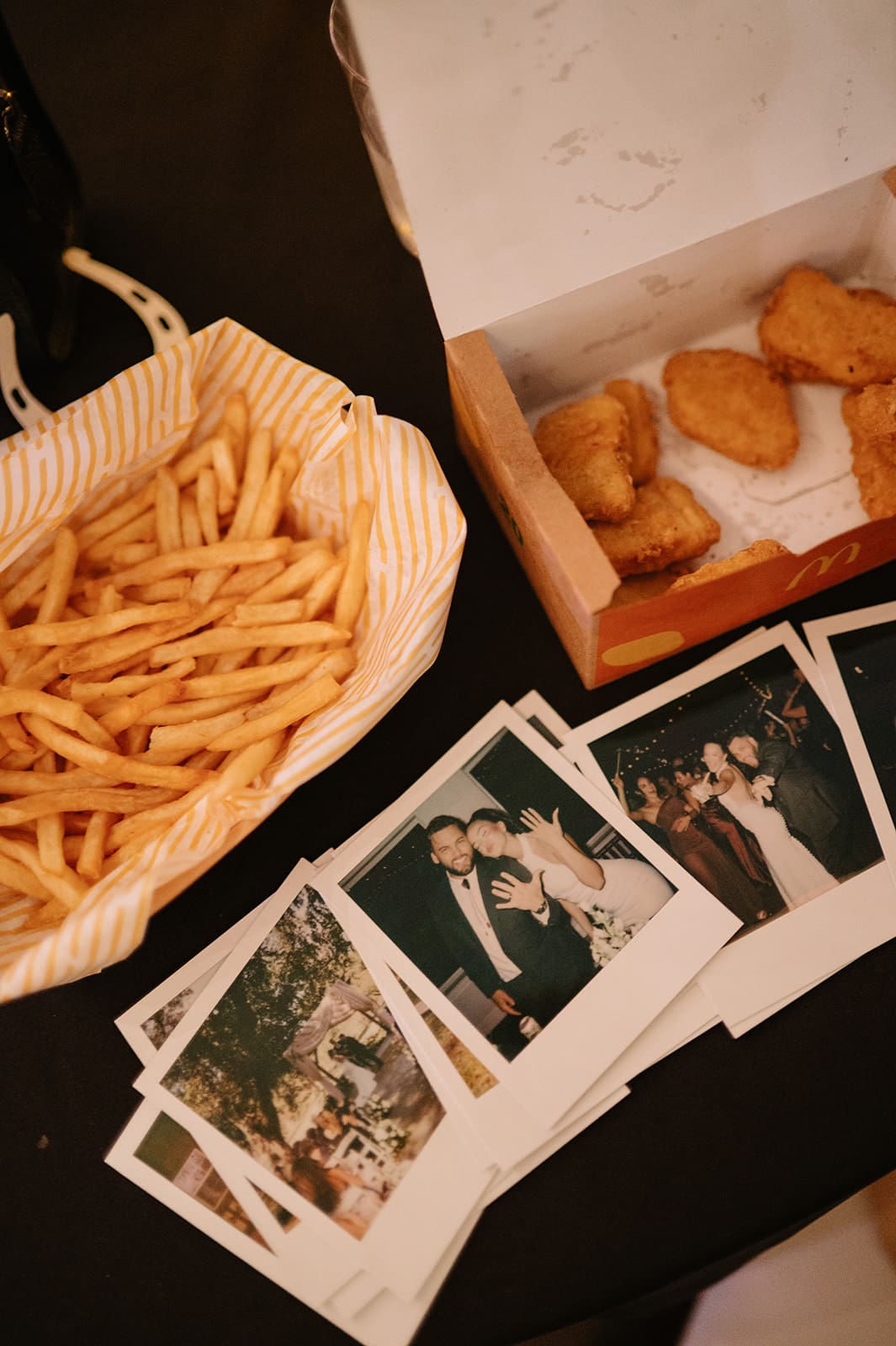 A striped bag of French fries, a box with chicken nuggets, and several instant photos spread out on a dark surface.