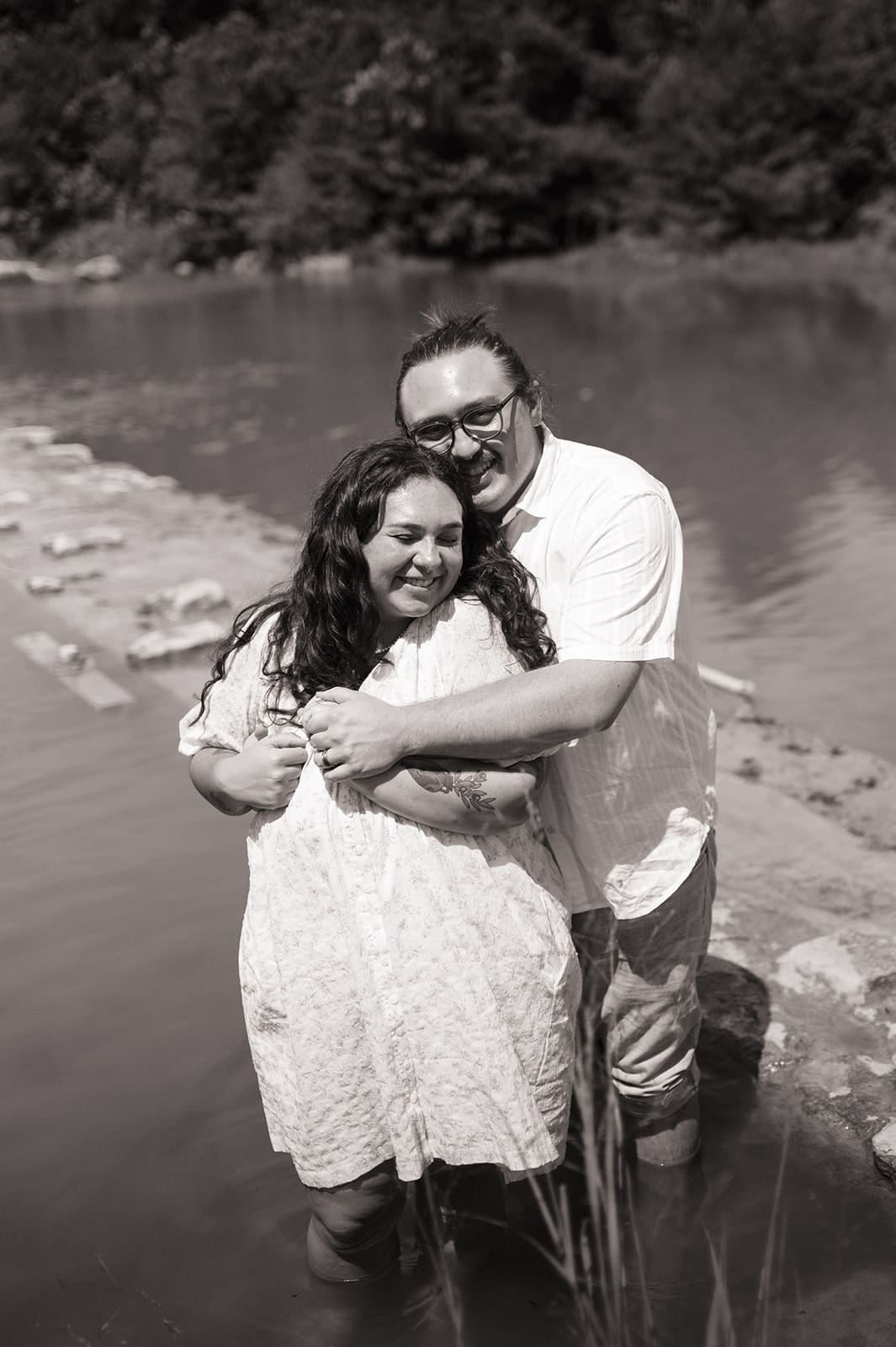 Two people stand in shallow water near a stone edge, holding hands and smiling at each other, with trees and a pond in the background.