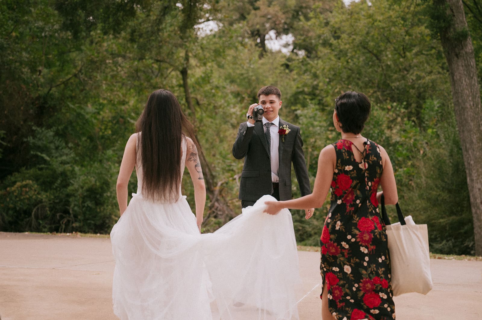 A person in a suit takes a photo of two women walking outdoors; one wears a white dress, the other a black floral dress, both seen from behind.