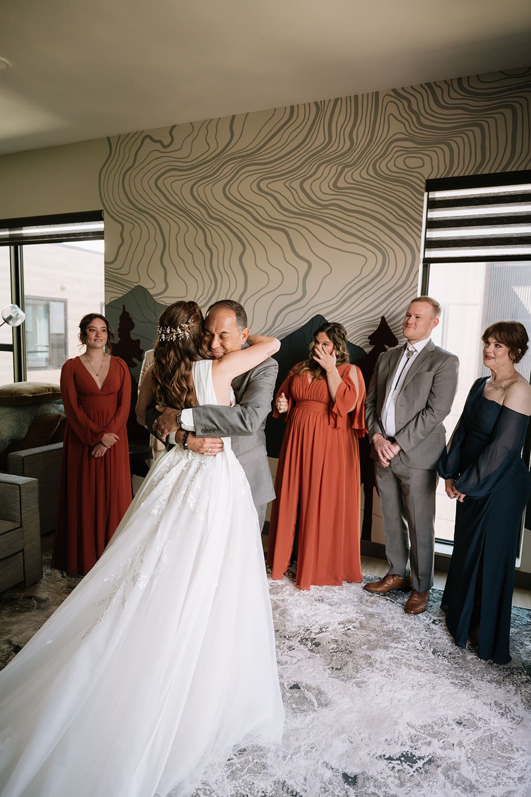 A bride in a white dress hugs a man in a suit while four adults look on, standing in a modern room with large windows and patterned walls.
