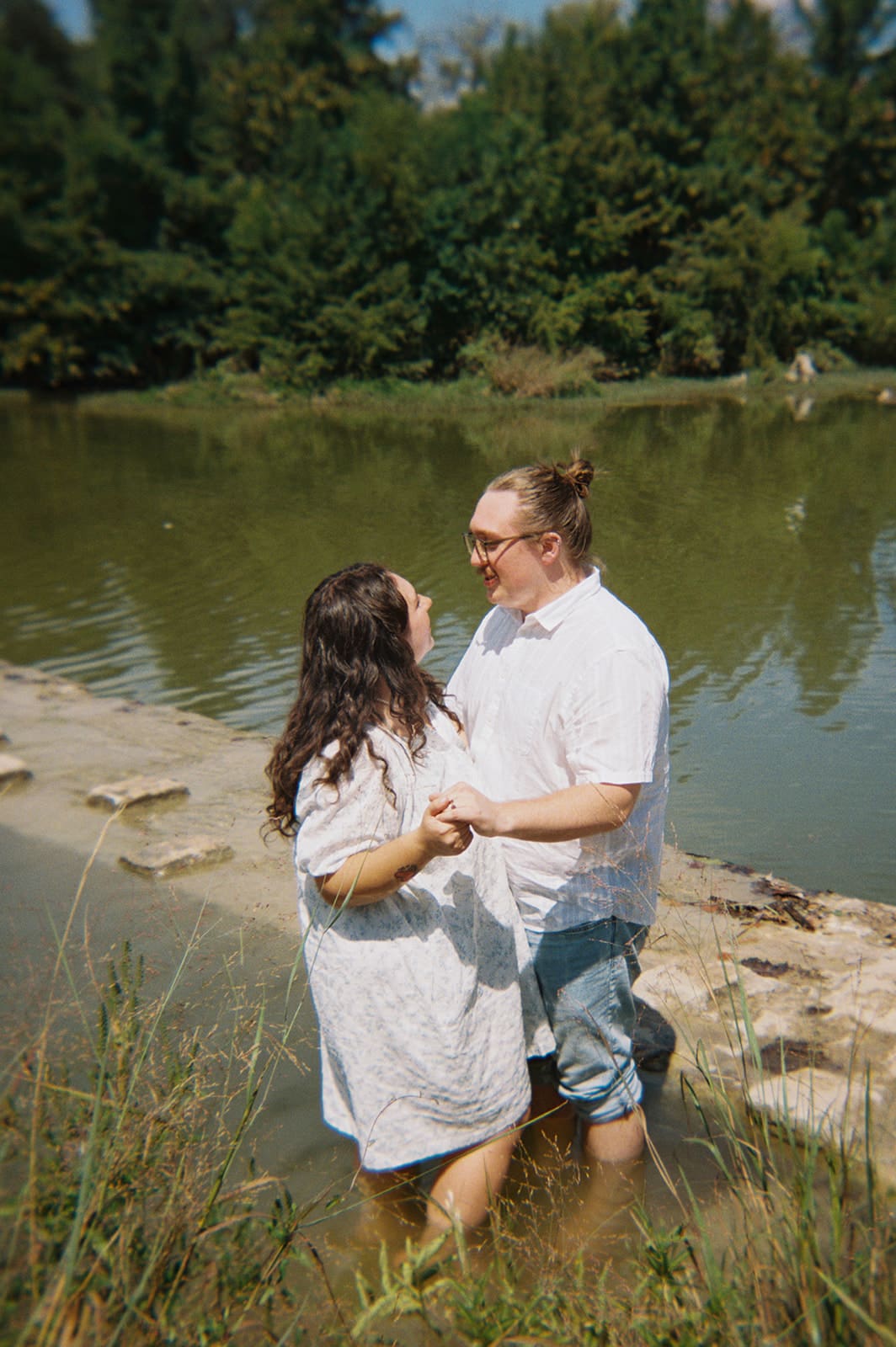 Two people stand in shallow water near a stone edge, holding hands and smiling at each other, with trees and a pond in the background.