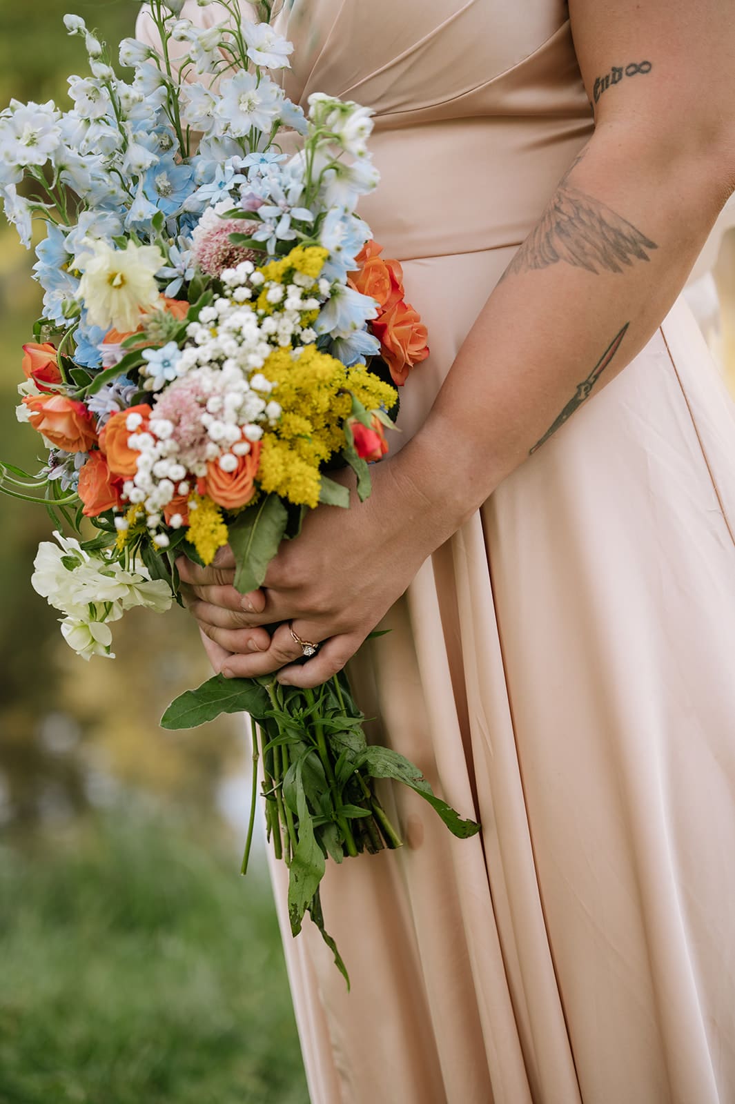 Person in a light pink dress holding a colorful bouquet of flowers, with a visible wing tattoo on their arm, standing outdoors.