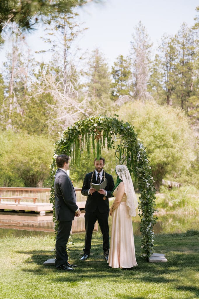A bride and groom stand under a flower-covered arch outdoors with an officiant, surrounded by trees and greenery during a wedding ceremony | 9 Wedding Venues in Oregon for an Intimate Celebration