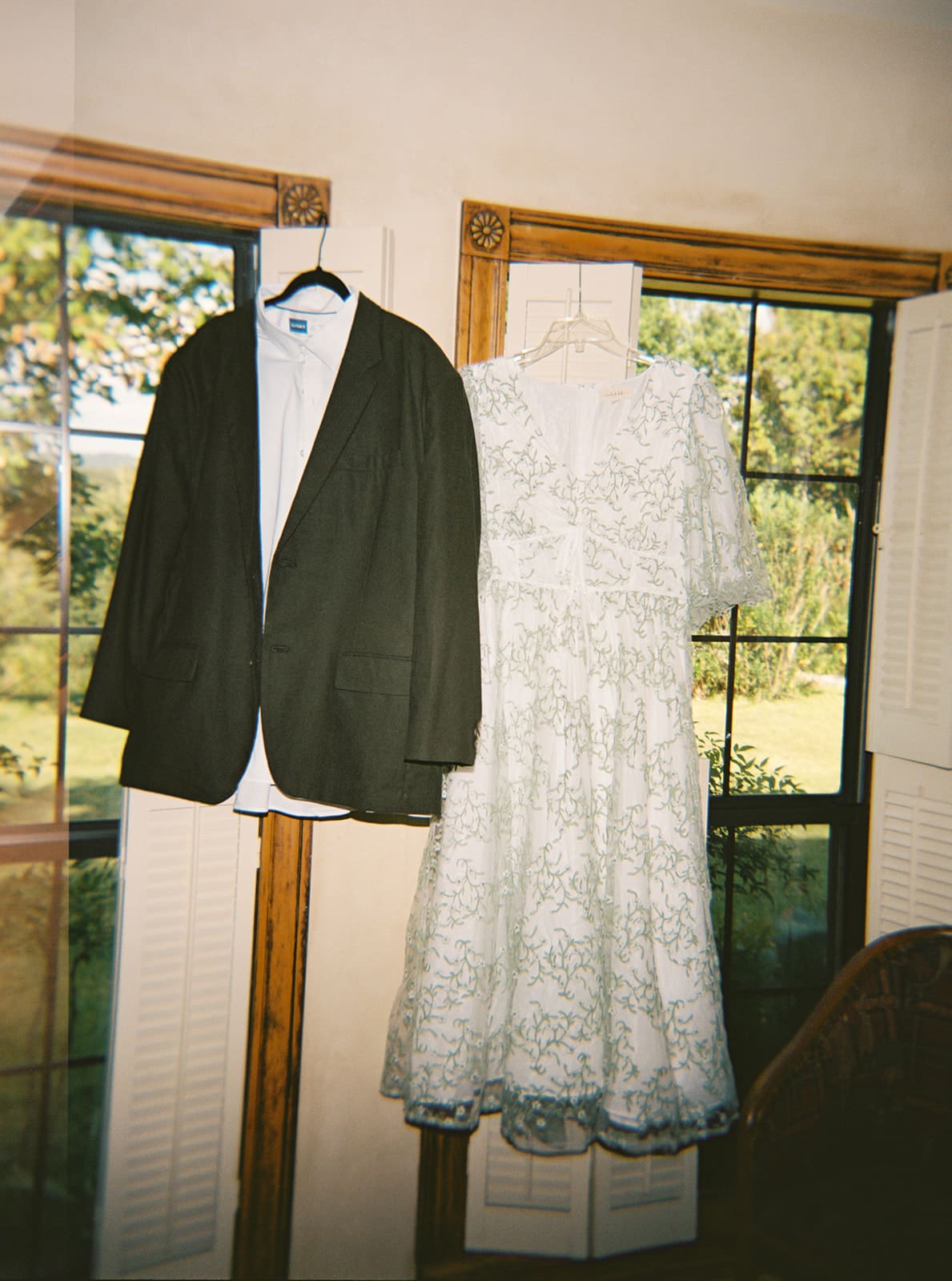 A black suit jacket and a white lace dress hang side by side on a wall between two windows with a view of greenery outside. | Unique Wedding Ideas