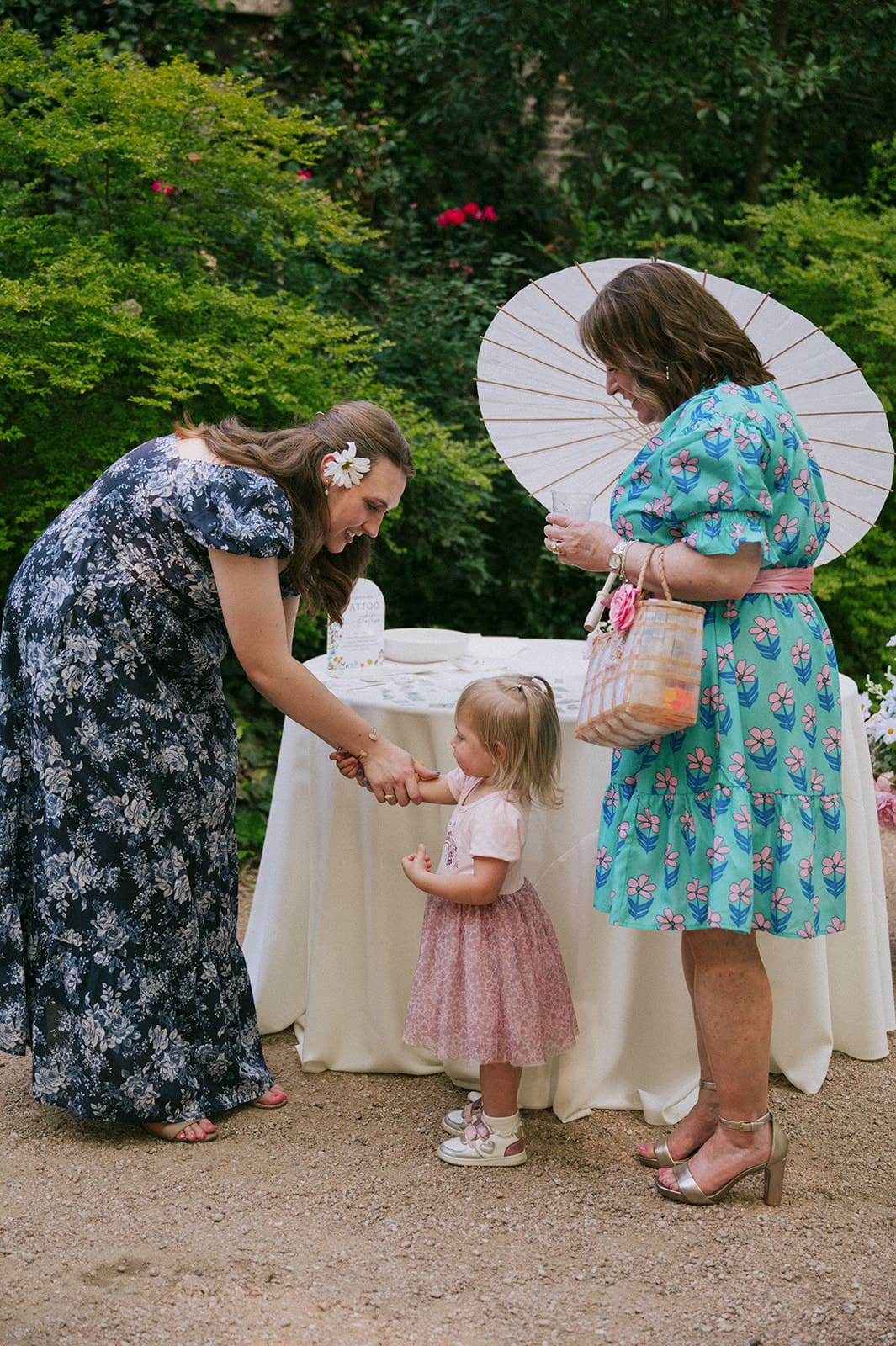 Two women and a young girl stand outdoors by a white table. One woman bends down to hand something to the girl. Green foliage and a parasol are in the background.