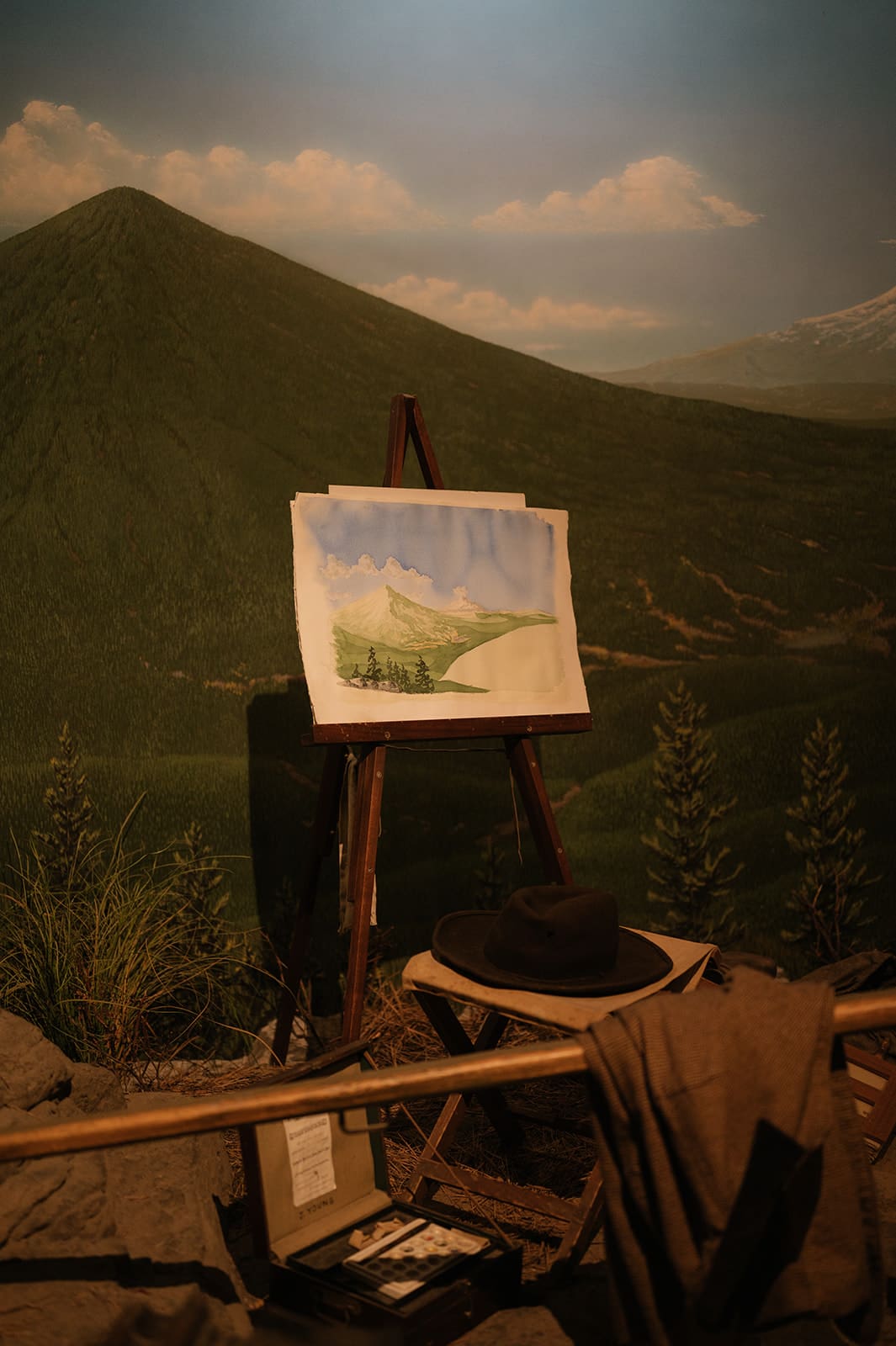 An easel with a landscape painting stands outdoors in front of a green mountain, surrounded by art supplies, a hat, and clothing.