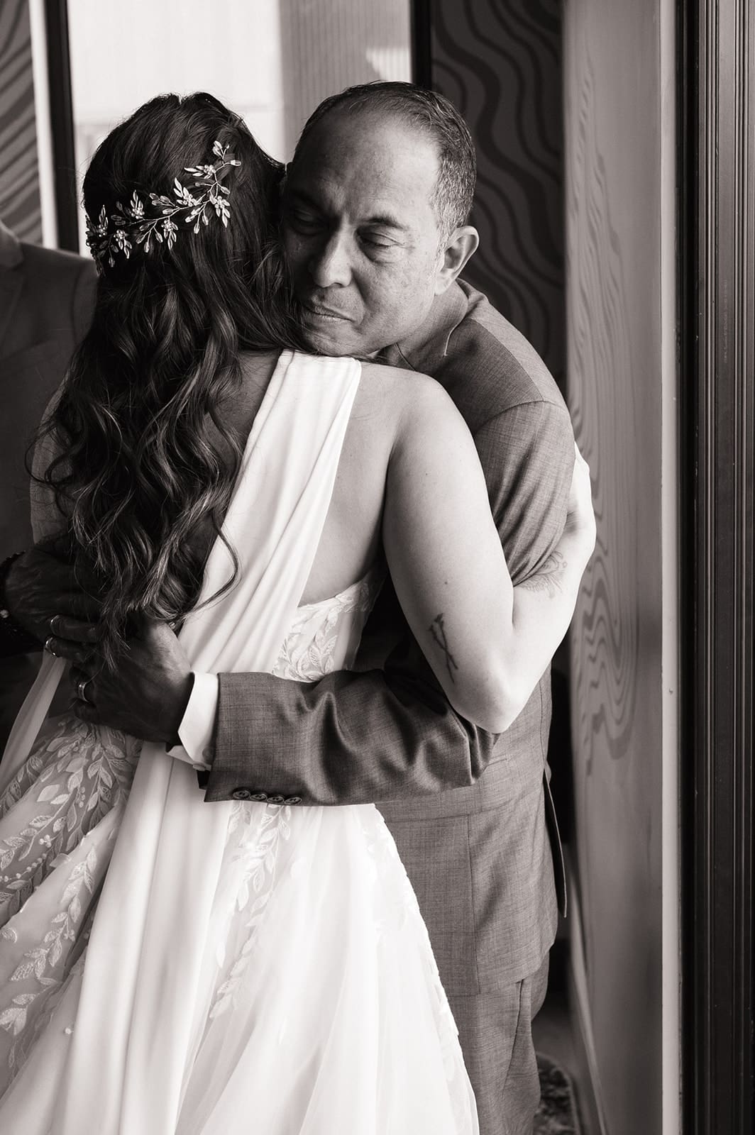 A bride in a white dress hugs a man in a suit while four adults look on, standing in a modern room with large windows and patterned walls.