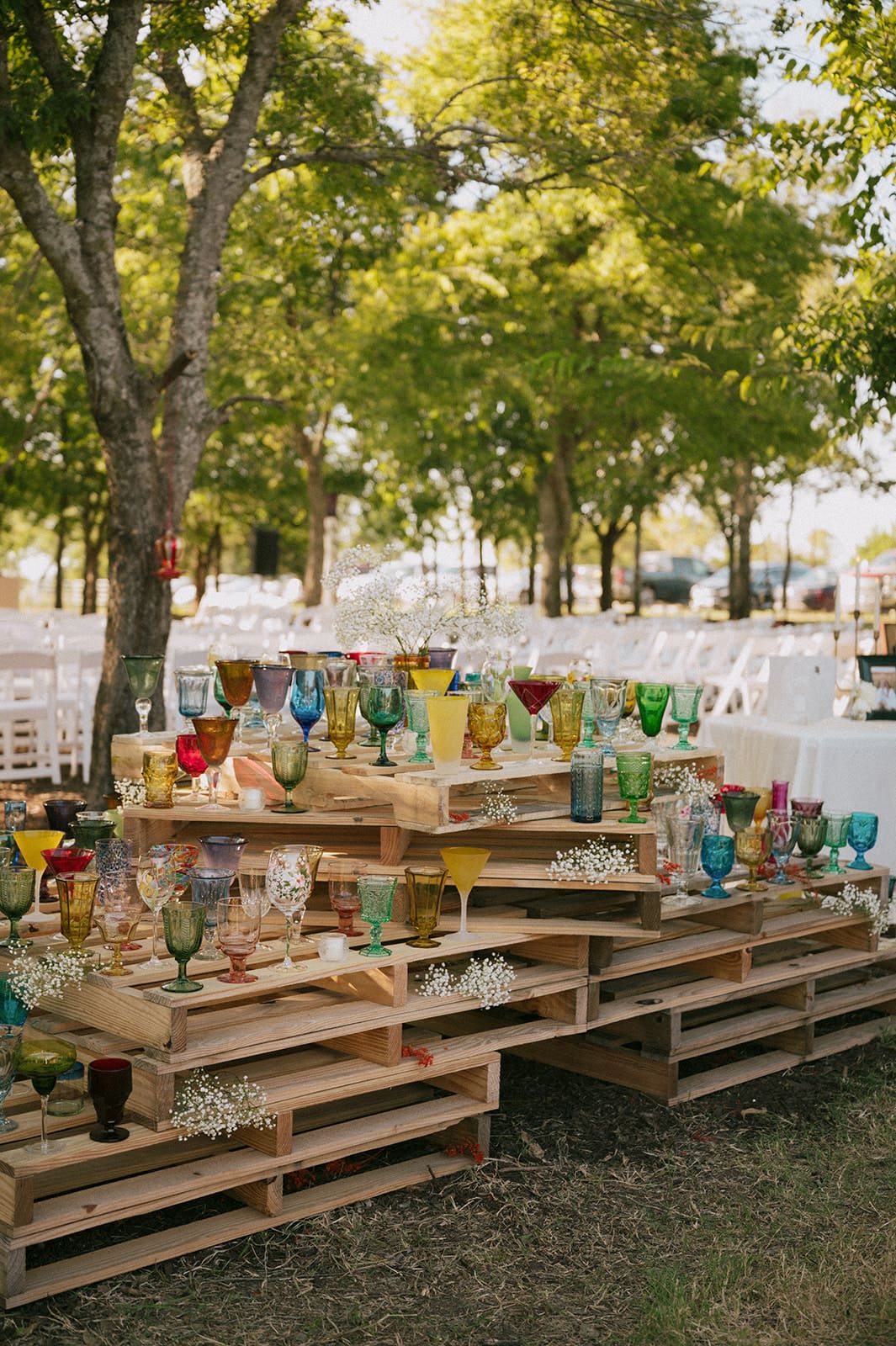 Colorful glass goblets and baby’s breath flowers are arranged on a wooden pallet table outdoors, with people and trees visible in the background.