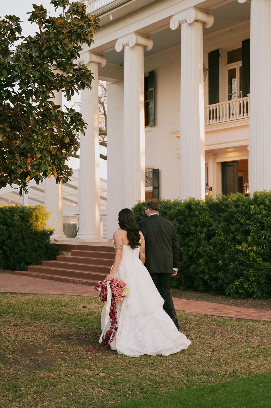 A bride and groom walk on grass toward a large white building with columns, holding hands and the bride holding a bouquet.