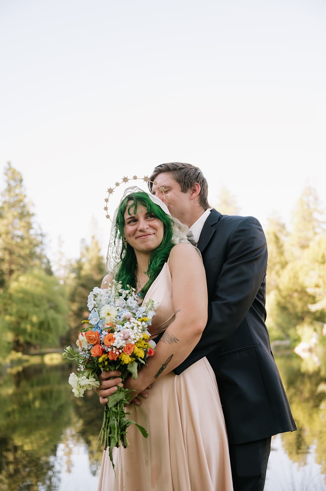 A couple poses outdoors by a lake; the woman in a light dress holds a bouquet and has green hair, while the man in a suit stands behind her, both looking happy.