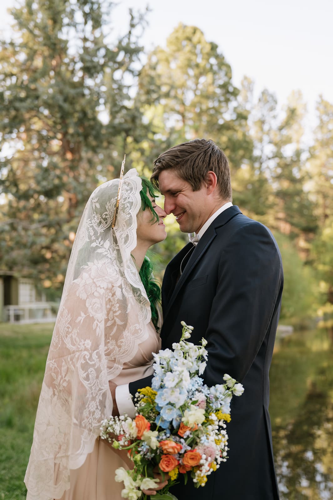 A bride and groom stand close together outdoors, smiling at each other. The bride holds a bouquet of flowers and wears a lace veil. Trees and a pond are visible in the background | 9 Wedding Venues in Oregon for an Intimate Celebration