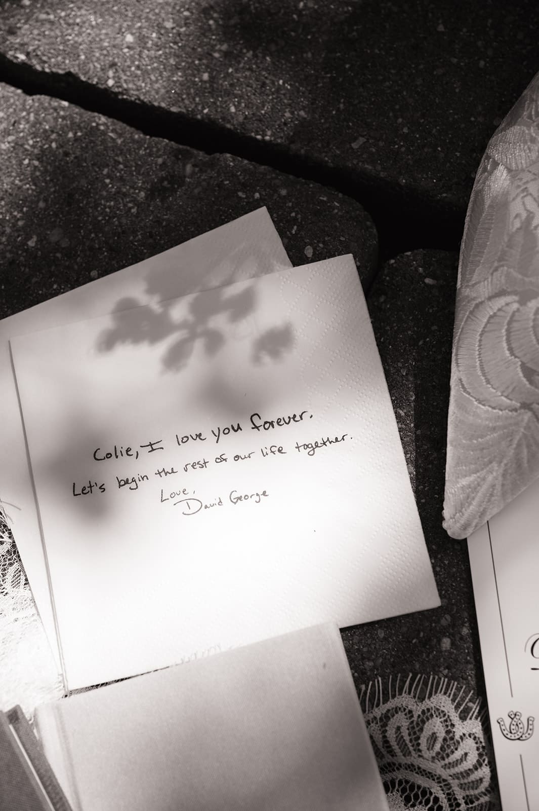 A flat lay of invitation cards, a green notebook, a small box, perfume bottles, a lace cloth, a ribboned pouch, and a photo on a stone surface with some foliage shadows.