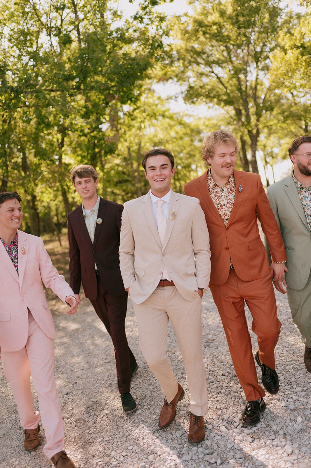 Five men in colorful suits walk together on a gravel path outdoors, surrounded by green trees and sunlight.