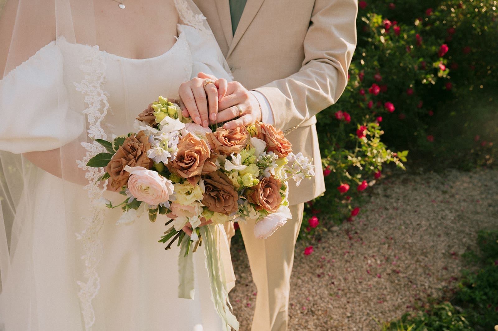 A bride in a white dress holds a bouquet of pastel and brown flowers, standing next to a person in a beige suit.
