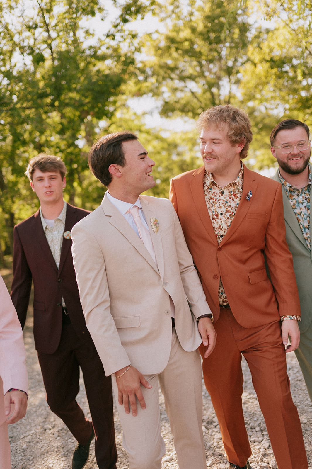 Five men in colorful suits walk together on a gravel path outdoors, surrounded by green trees and sunlight.